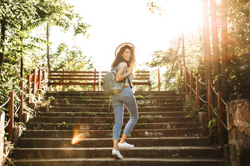 Obraz premium Photo of beautiful teenage girl 18-20 wearing summer clothes and straw hat, walking up stairs in green park on sunny day