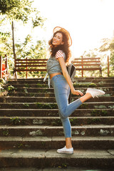 Obraz premium Full length image of lovely curly woman 18-20 wearing summer clothes and straw hat, smiling and standing on stairs in green park on sunny day