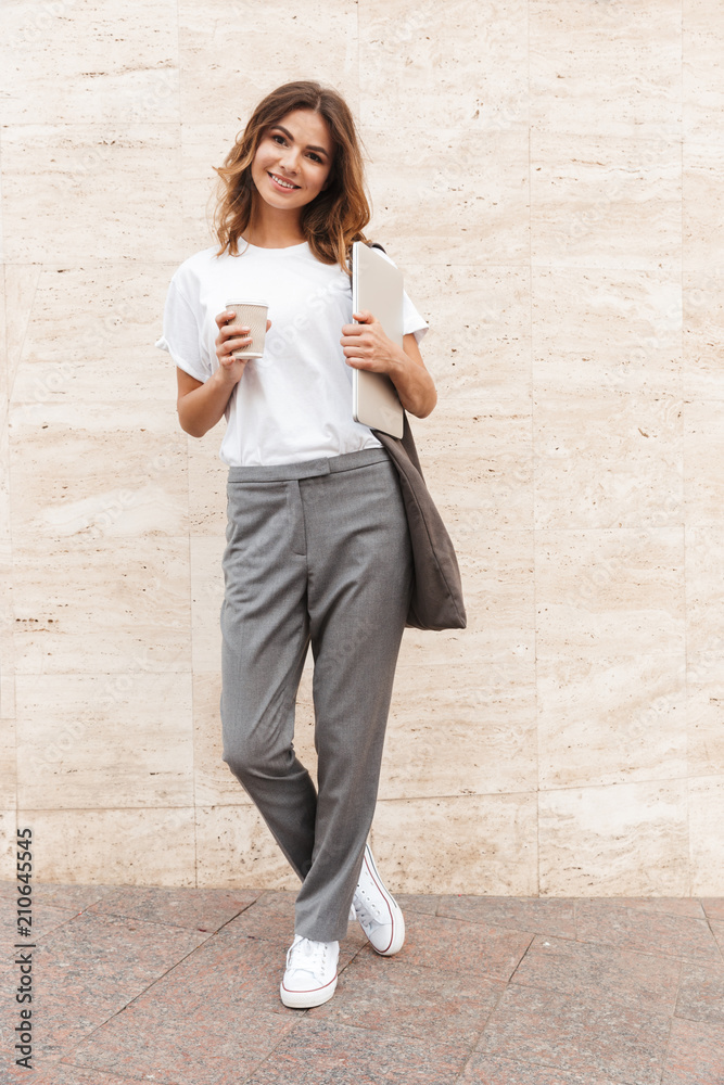 Poster Full length image of beautiful modern woman standing against beige wall outdoor with silver laptop, and takeaway coffee in hands