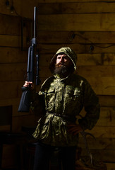 Man with beard wears camouflage hooded clothing, wooden interior background. Huntsman concept. Hunter, brutal hipster with gun prepare rifle for hunting. Macho on strict face at gamekeepers house.
