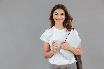 Photo of beautiful young woman smiling and standing against gray background isolated with silver laptop, and takeaway coffee in hands