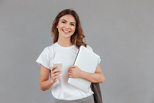 Image of beautiful young woman smiling and standing against gray wall isolated with silver laptop, and takeaway coffee in hands