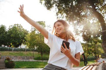 Image of smiling friendly woman sitting on bench in green park on summer day, and waving hand aside...