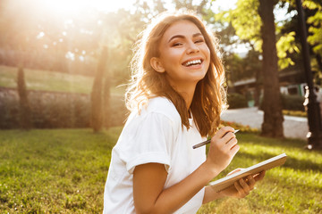 Obraz premium Zdjęcie radosnej kobiety po dwudziestce z szerokim uśmiechem siedzącej na zielonej trawie w parku ze skrzyżowanymi nogami w letni dzień i zapisującej notatki piórem
