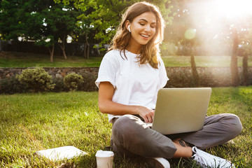 Portrait of pretty young woman sitting on green grass in park with legs crossed during summer day, while using laptop and wireless earphone for video call - Powered by Adobe