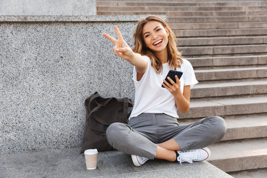 European smiling woman sitting on street stairs with legs crossed on summer day, and showing peace sign with mobile phone in hand