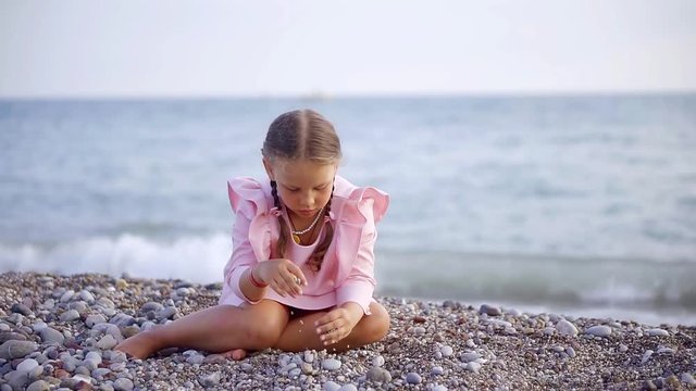 alone pretty little girl is wearing nice pink dress is sitting on a pebble beach near sea and playing