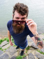 Man with beard and mustache wears sunglasses, water surface on background. Hipster on winking face...