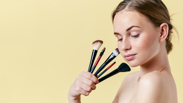 Pretty redhead teenage girl with freckles, looking down, holding diverse make up brushes. Model with light nude make-up. Studio beauty portrait on yellow background.