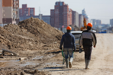 Two workers in helmets drive a wheelbarrow against the background of new buildings.
