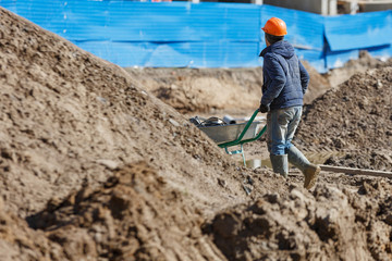 A worker in a helmet carries a wheelbarrow against the background of construction debris.