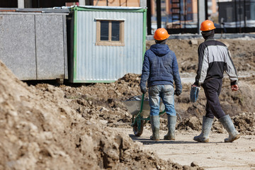 Two workers in helmets are driven a wheelbarrow on the background of a building shed.