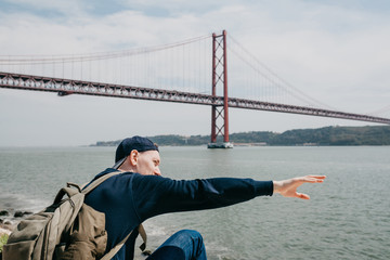 Naklejka premium A young traveler or a tourist with a backpack on the waterfront in Lisbon in Portugal next to the 25th of April Bridge