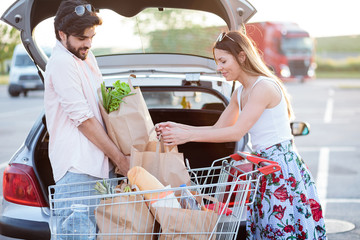 Happy young couple loading grocery bags into a car trunk at a parking lot in front of a shopping mall. Boyfriend lifts heavy bag while girlfriend is helping. © Ivan