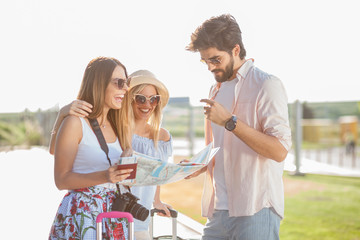 Young man showing directions on the map to two beautiful young female tourists. Standing in front of the airport terminal building with suitcases, maps, passports and camera.