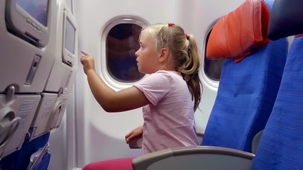 little child girl is passenger of airplane, is sitting on a seat in cabin, looking on a display in chair