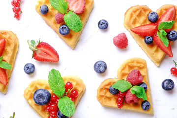 Heart shape belgium waffles with assorted berries mix, strawberry, blueberry, raspberry and red currant decorated with mint leaf on white ceramic plate. Close up, copy space, background, top view.