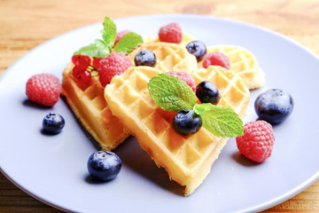 Heart shape belgium waffles with assorted berries mix, strawberry, blueberry, raspberry and red currant decorated with mint leaf on white ceramic plate. Close up, copy space, background, top view.