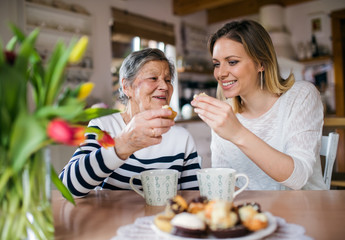 An elderly grandmother with an adult granddaughter sitting at the table at home, eating cakes. - obrazy, fototapety, plakaty