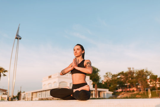 Photo of thoughtful lady sitting in lotus pose and dreamily looking aside. Young pretty woman in black sporty top and leggings meditating while practicing yoga