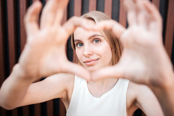 Naklejka premium Portrait of a girl making a heart from her fingers and showing it on camera. Also she is looking at it and smiling. Isolated on striped background.