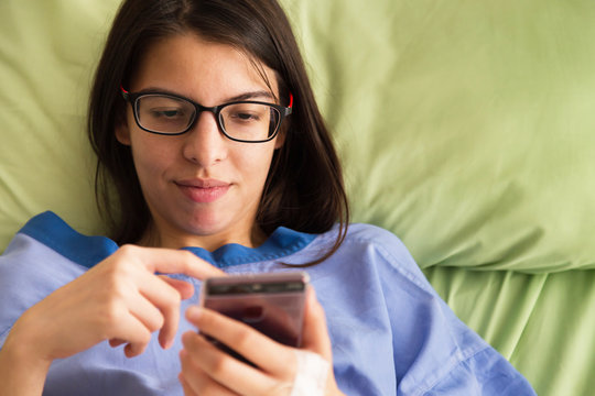 Beautiful Young Patient Women Lying In  Hospital Bed And Smiling With Smart Mobile Phone While Recovering In Hospital. Recovering Patient Women Are Happy Working On Business Mobile Phone In Hospital.