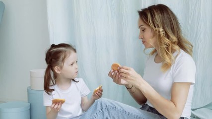 Mom and little girl feed each other cookies. A woman with a little daughter are sitting on the floor. A young mother with a little daughter tasting cookies and showing the likes.