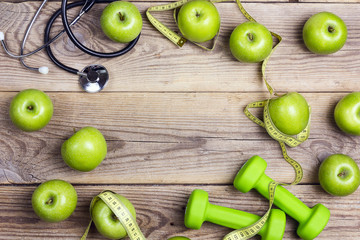 Fototapeta na wymiar Green dumbbells and apples with measuring tape and stethoscope on wooden background.