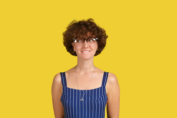 Studio portrait of mysterious charming young European female with shaggy hairstyle looking sideways with enigmatic smile. Emotional beautiful lovely woman conceived a surprise. Yellow wall background