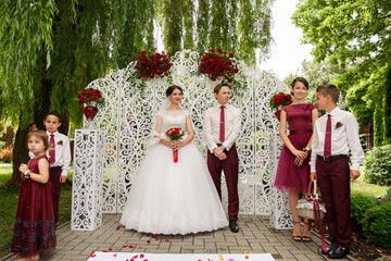 Naklejka premium Young gorgeous bride and handsome groom standing near the wedding ornament wall, with bridesmaid, groomsman and kids, wedding ceremony. At the park. Marsala style.