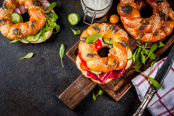 Variety of homemade bagels sandwiches with sesame and poppy seeds, cream cheese,  ham, radish, arugula, cherry tomatoes, cucumbers, with ingredients on dark concrete background copy space top view
