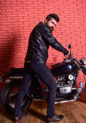 Fototapeta na wymiar Man with beard, biker in leather jacket near motor bike in garage, brick wall background. Hipster, brutal biker on serious face in leather jacket sits down on motorcycle. Masculine hobby concept.