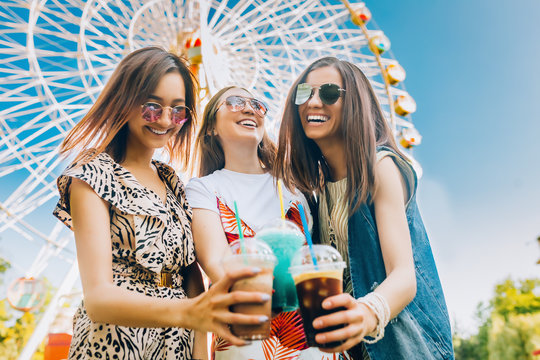 Summer lifestyle portrait multiracial women enjoy nice day, holding glasses of milkshakes. Happy friends inin front of ferris wheel. Best friends girls having fun, joy. Lifestyle. Asian, jewess and