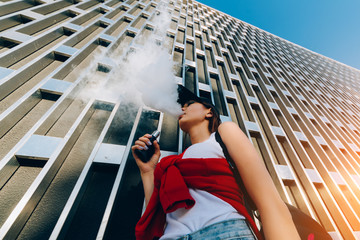 Pretty young hipster asian woman in black hat vape ecig, vaping device at the sunset. Toned image.