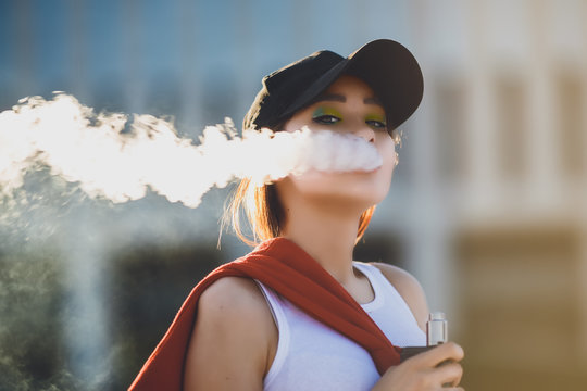 Pretty young hipster asian woman in black hat vape ecig, vaping device at the sunset. Toned image. Closeup.