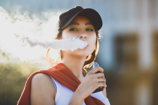 Pretty young hipster asian woman in black hat vape ecig, vaping device at the sunset. Toned image. Closeup.