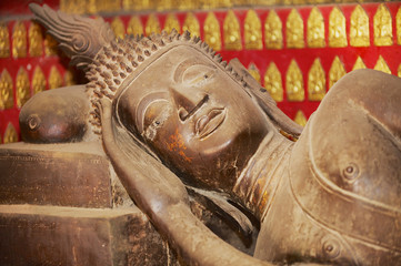 Reclining Buddha statue in a red chapel with many small Buddha statues at the background in Wat Xieng Thong temple in Luang Prabang, Laos.