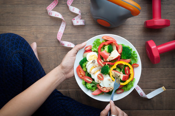 Sporty woman holding salad dish with sport equipments on wooden floor, Top view. Healthy and wellbeing concept