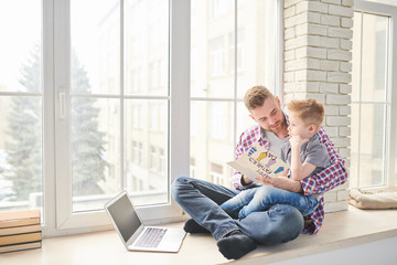 Full length portrait of modern young man embracing his cute son while sitting on window together and reading card for Fathers day, copy space