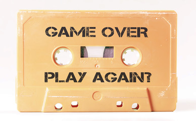 A vintage cassette tape from the 1980s era (obsolete music technology) with the text Game Over and Play Again. Color: cream, sand. White background.
