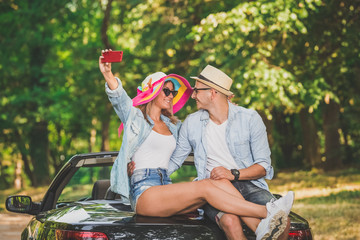 Fashionable couple sitting on the cabriolet trunk and taking self portrait.