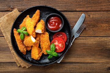 Delicious crispy fried breaded chicken breast strips with ketchup on black plate.
