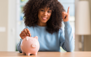 African american woman saves money in piggy bank surprised with an idea or question pointing finger...