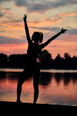 Fototapeta premium Sporty woman posing near city lake on sunset. Fitness, yoga, crossfit exercises. Fit, strong bodies, healthy lifestyle. Outdoors workout on fresh air. Colorful sky view. Water reflection.