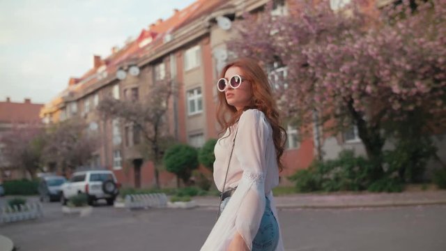 Young beautiful fashionable woman wearing stylish round sunglasses, white chiffon blouse, wrist watch, with small bag posing in street of city