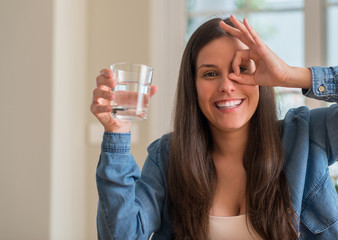 Young woman drinking glass of water at home with happy face smiling doing ok sign with hand on eye looking through fingers