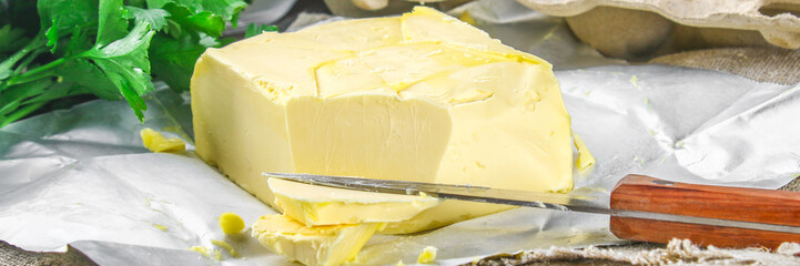 A bar of butter is cut into pieces on a wooden board with a knife, surrounded by milk, eggs and parsley on a brown table. Ingredients for cooking.