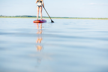Fototapeta premium Young woman in black swimsuit paddleboarding on the lake with calm water and beautiful reflection during the sunset. Cropped image with no face