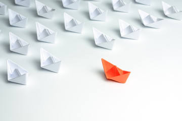 Leadership concept with orange paper ship standing out from the group of white ships on white...