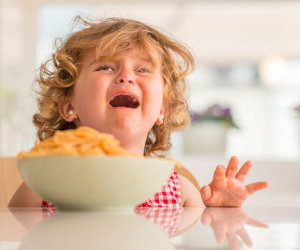Beautiful blond child eating spaghetti with hands crying with tantrum at home.
